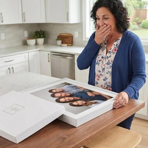 May include: A white rectangular gift box with a lid featuring a small logo, containing a framed photograph of four people stacked vertically. The box is resting on a wooden surface, showcasing a high-quality print of a family portrait inside.