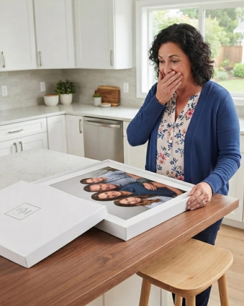 May include: A white rectangular gift box with a lid featuring a small logo, containing a framed photograph of four people stacked vertically. The box is resting on a wooden surface, showcasing a high-quality print of a family portrait inside.
