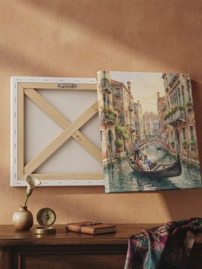 以下が含まれることがあります： ゴンドラ、建物、橋が描かれたヴェネツィアの運河の絵画がプリントされた正方形のキャンバス。キャンバスは裏側から見えており、木製のストレッチャーフレームと、上部のバーに取り付けられた金属製の吊り下げ金具が確認できます。