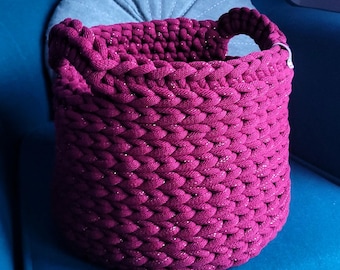 May include: A round, burgundy crochet basket with a handle. The basket is made of thick, textured yarn with a subtle shimmer. It sits on a blue chair with a gray pillow behind it. The basket is designed for storage.