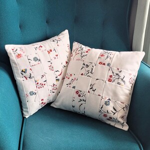 May include: Two square cushions with a patchwork design. The cushions have a white background with floral patterns in red, blue, and pink. They are placed on a teal armchair.