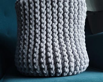 May include: A gray, cylindrical, hand-knitted basket with a textured, rope-like appearance. The basket is made of thick, soft yarn and sits on a teal surface. The basket is a decorative storage item.