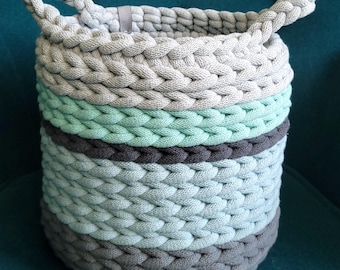 May include: A round, hand-knitted basket with two handles. The basket is made of thick, braided yarn in shades of gray, mint green, and blue. The basket is sitting on a teal surface.
