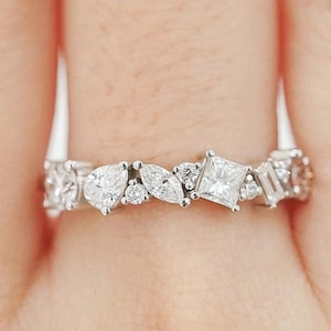 May include: A white gold ring with a variety of gemstones. The ring showcases heart, marquise, square, and baguette-cut stones, creating a distinctive design. The gemstones are set closely together, forming a continuous band. The ring is a piece of jewellery.