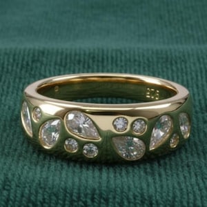 May include: A gold band ring with a series of pear-shaped and round clear gemstones. The ring is set against a dark green textured background. The ring's design features a repeating pattern of gemstones.