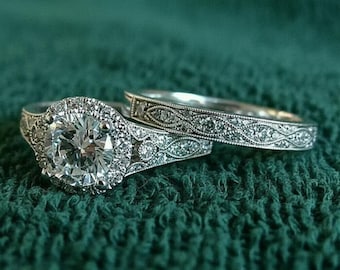 May include: A diamond engagement ring set with a large, round-cut center stone and a halo of smaller diamonds. The band features intricate detailing. A matching wedding band with similar detailing is also present. The rings are set against a green textured background.
