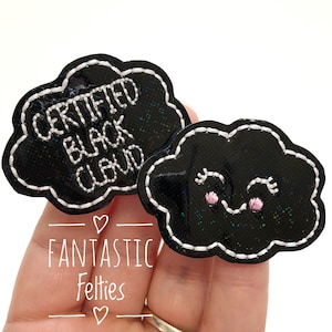 May include: Two black cloud-shaped patches with white stitching. One patch has the text "CERTIFIED BLACK CLOUD" embroidered in white. The other cloud has a smiling face with pink cheeks and eyelashes. The patches are glittery.
