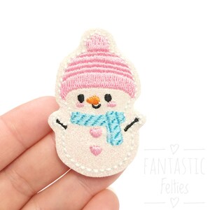 May include: A pink and white felt snowman applique with a blue scarf and a pink hat. The snowman has a smiling face with black eyes and a carrot nose.