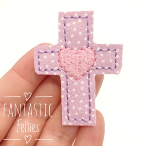 May include: A pink felt cross with white polka dots and a pink felt heart in the center. The cross is stitched with purple thread.