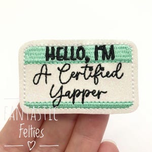 May include: A rectangular embroidered patch with a white glittery background and mint green borders. The black embroidered text reads: HELLO, I'M A Certified Yapper. The patch features rounded corners and a stitched edge finish.