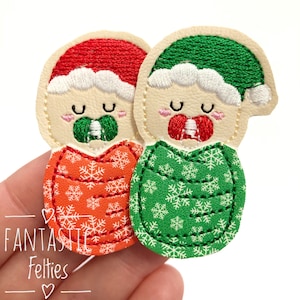 May include: Two embroidered felt appliques shaped like babies wearing holiday hats. One baby wears a red hat and orange snowflake swaddle with a green pacifier. The other wears a green hat and green snowflake swaddle with a red pacifier.