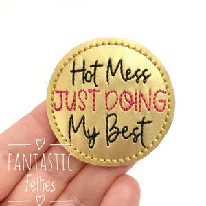 May include: A gold round felt patch with black and pink embroidery that reads "Hot Mess Just Doing My Best".