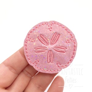 May include: Pink felt sand dollar with a glittery finish. The sand dollar is embroidered with a detailed design.