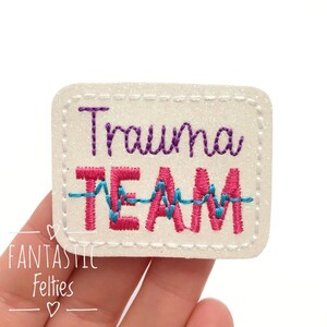 May include: White glitter iron-on patch with pink and blue embroidery. The patch says "Trauma Team" with a blue embroidered EKG line.