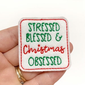 May include: White felt square patch with red and green embroidery. The text reads "Stressed Blessed & Christmas Obsessed".