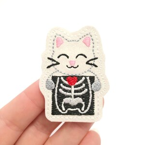 May include: An embroidered patch featuring a smiling white cat with pink ears and black whiskers. The cat is holding a black and white x-ray of a skeleton with a red heart. The patch has a stitched border.