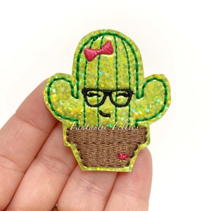 May include: A glittery green cactus-shaped felt applique with a smiling face, black glasses, and a pink bow. The cactus is in a brown pot. The text "Fantastic Felties" is at the bottom. This item is likely for crafting or embellishment.