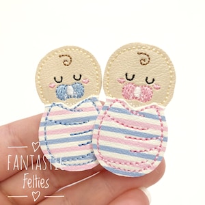 May include: Two embroidered felt baby appliques featuring swaddled infants with closed eyes and rosy cheeks. One baby has a blue pacifier and blue-striped swaddle, while the other has a pink pacifier and pink-striped swaddle. Text reads FANTASTIC FELTIES.
