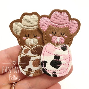 May include: Two felt cowgirl appliques with pink and brown cow print patterns. The appliques are wearing pink and beige cowboy hats with embroidered details. The appliques have embroidered eyes and noses.