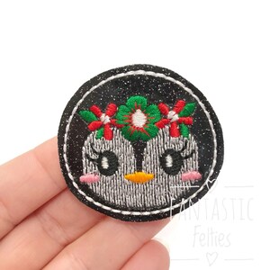 May include: Embroidered patch featuring a cute penguin with a floral crown. The penguin has a black and white body, pink cheeks, and an orange beak. The patch is round with a black glitter background and white stitching.