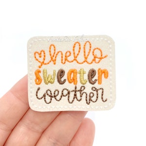 May include: A white felt patch with brown stitching around the edges. The patch has the text "hello sweater weather" embroidered in orange, brown, and yellow thread.