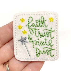 May include: Green glitter iron-on patch with the text "faith & trust pixie dust" in white with a silver glitter star and wand. The patch is surrounded by a white stitched border and four yellow stars.