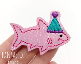 Puede incluir: Aplique de fieltro rosa con forma de tiburón con gorro de fiesta. El aplique tiene una textura brillante y está contorneado con costuras rosas. El gorro de fiesta es turquesa con un pompón morado. Texto "FANTASTIC Felties" en la parte inferior.