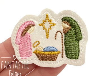 May include: A white glittery felt patch featuring an embroidered nativity scene. The design includes a pink-clad figure, a green-clad figure with a brown staff, a central yellow star, and a baby in a brown manger with a blue blanket.