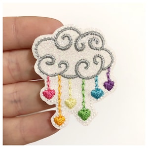 May include: A white, glittery cloud-shaped patch with silver embroidered outlines. The cloud has rainbow-colored heart-shaped raindrops. The text "Fantastic Felts" is visible on the cloud.