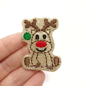 May include: Gold glitter reindeer applique with a red nose and green ornament. The reindeer is sitting with its legs crossed and has a brown outline.