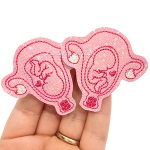 May include: Two pink felt appliques with embroidered details depicting a fetus inside a womb. The appliques have a glittery finish and are held in a hand. The text "Fantastic Felties" is visible.