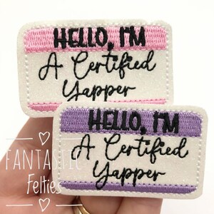 May include: Two rectangular embroidered patches featuring a white glittery background with pink or purple borders. The text reads "HELLO, I'M A Certified Yapper" in black embroidery. The patches have a stitched edge and a name tag design.
