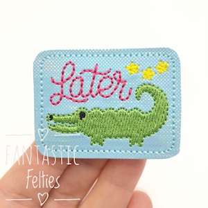 May include: A rectangular light blue patch with a subtle white dot pattern, featuring an embroidered green alligator, three small yellow stars, and the word "Later" stitched in pink cursive thread with a decorative border stitch.