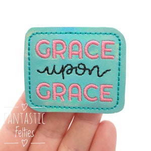 May include: A teal rectangular patch with the words "GRACE upon GRACE" embroidered in pink and black thread. The patch has a stitched border and a holographic sheen. The patch is a feltie.