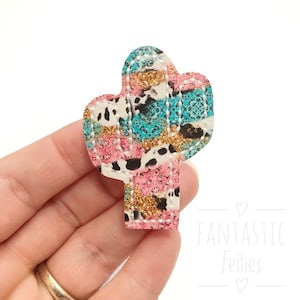May include: A pink, turquoise, and white cactus-shaped felt applique with a cow print and paisley pattern. The felt applique has a glittery finish and is stitched around the edges.