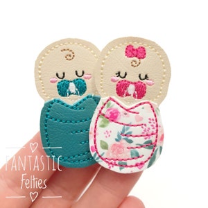 May include: Two felt baby dolls, one in a teal swaddle and the other in a pink floral swaddle. Both dolls have embroidered faces and pacifiers.