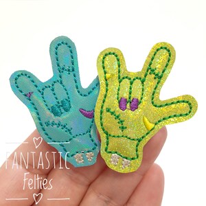 May include: Two felt hand patches in the shape of the American Sign Language symbol for "I love you". The left hand is turquoise blue with purple stitching and the right hand is yellow-green with purple and yellow stitching.