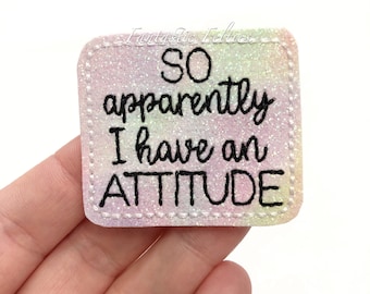 May include: A square-shaped, glittery, rainbow-colored patch with white stitching around the edges. The patch has black text that reads "So apparently I have an ATTITUDE".