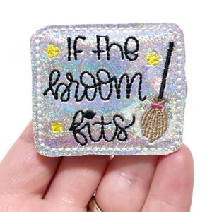 May include: A glittery, square-shaped patch with the phrase "If the broom fits" in black script. A small broom with a pink handle is on the right. Yellow star accents are scattered around the text. The patch is held in a hand.