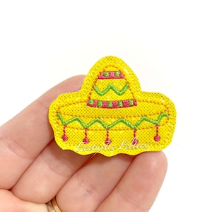 May include: A yellow sombrero-shaped felt applique with green and pink embroidered details. The applique has a dotted texture and the text "Fantastic Felties" is visible.