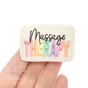May include: A rectangular, off-white felt patch embroidered with the words "Massage Therapy" in black and rainbow-colored thread. The patch has a stitched border and is held in a hand.
