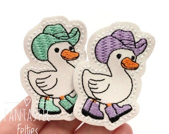 May include: Two embroidered duck patches. One duck wears a green cowboy hat and boots, the other a purple hat and boots. Both ducks have orange beaks and are outlined in black stitching. The patches are white with a white stitched border. The text "Fantastic Felties" is visible.