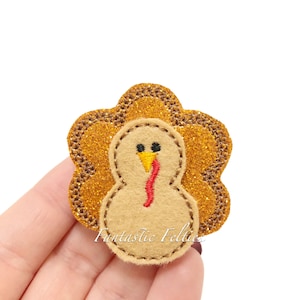 May include: A turkey-shaped felt applique featuring a tan felt body with black embroidered eyes, a yellow beak, and a red wattle. The turkey is layered over a gold glittery scalloped background with brown stitched edges and the text Fantastic Felties.