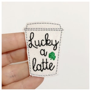 May include: White glitter coffee cup with black embroidery that says "Lucky a latte" and a green shamrock.