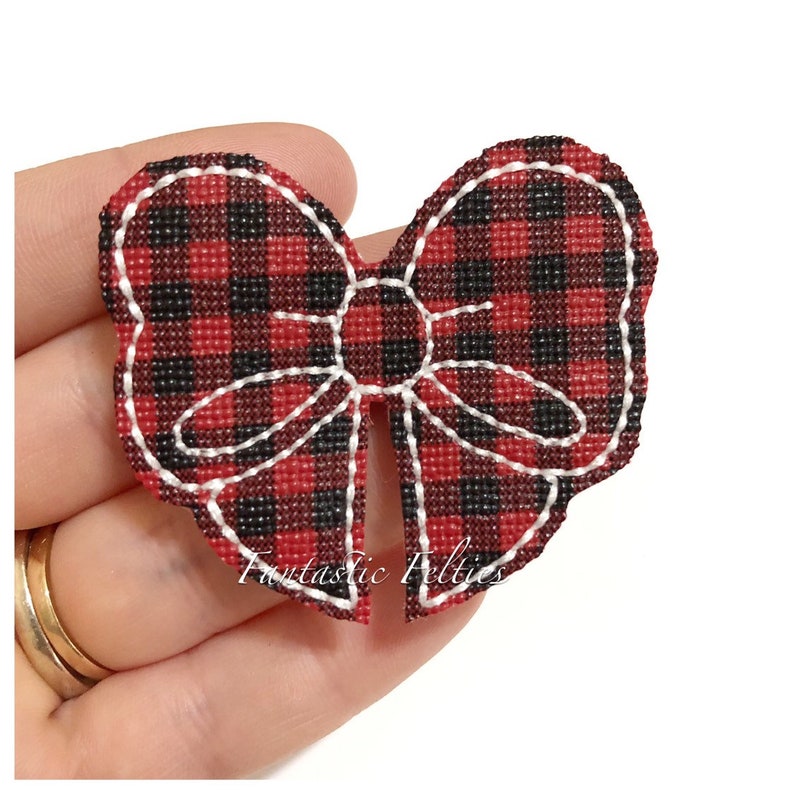 May include: A red and black plaid bow-shaped felt applique with white stitching. The bow has a scalloped edge and the text "Fantastic Felties" is printed on the bottom.