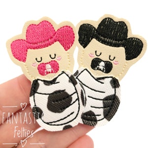 May include: Two felt cowboy baby appliques, one with a pink hat and the other with a black hat. Both are wearing a black and white cow print blanket.