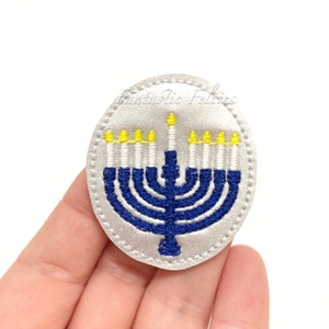 May include: A white oval felt patch with a blue menorah embroidered on it. The menorah has nine candles, with yellow flames. The text "Fantastic Felties" is embroidered in the top of the patch.