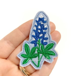 May include: Bluebonnet flower embroidered patch with green leaves. The patch is on a light blue felt background.