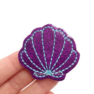 May include: A purple glitter seashell-shaped felt applique with light blue stitching. The applique has a scalloped edge and the text "Fantastic Felties" is visible on the bottom. This item is likely used for crafting or embellishment.