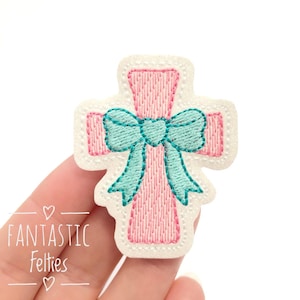 May include: A pink embroidered cross applique with a teal bow. The cross is outlined in white and has a stitched texture. The text "Fantastic Felties" is below the cross.